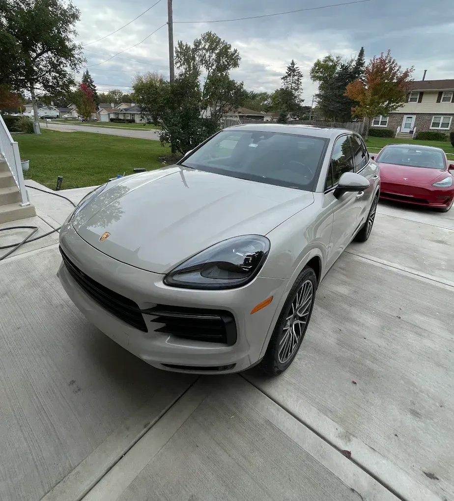 Silver Porsche SUV parked in a driveway with a red sports car behind it under cloudy skies