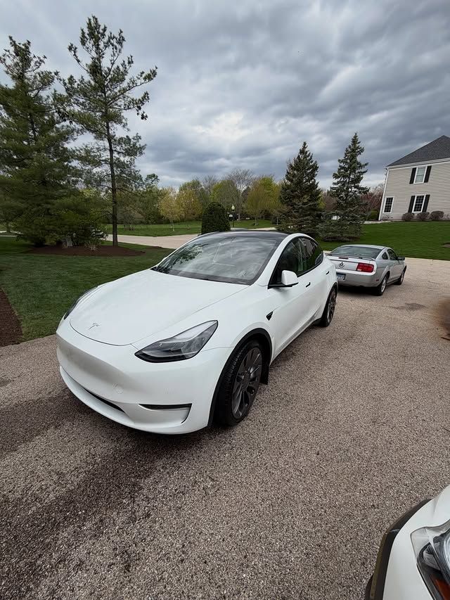 White Tesla parked on a gravel driveway with trees, a silver car, and a house under cloudy skies