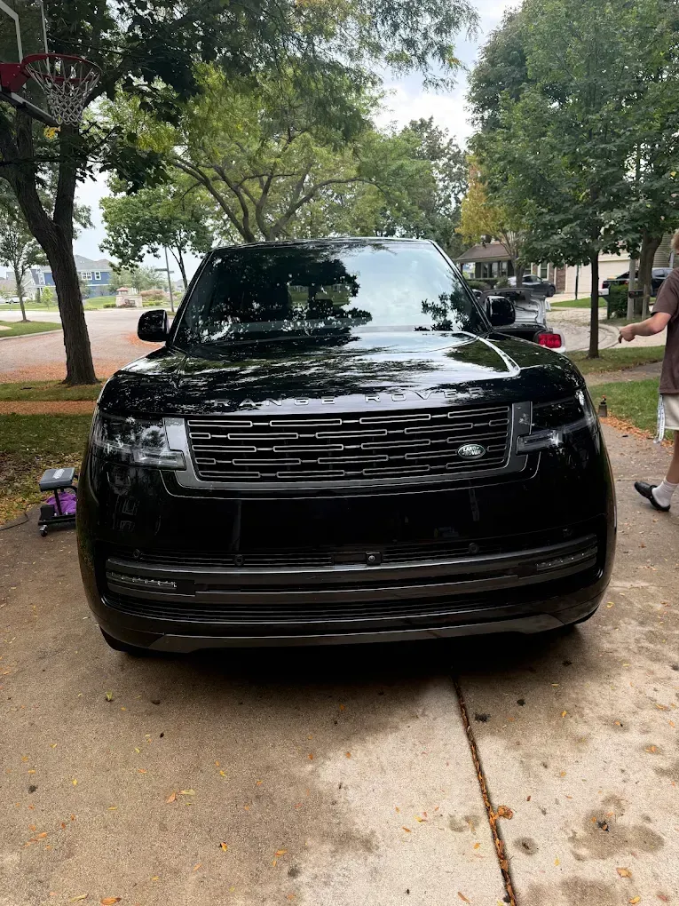 Black SUV parked on a leafy driveway, front view with trees and sidewalk in the background