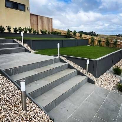 Grey stone steps and patio lead to a grassy yard with dark grey retaining walls and outdoor lights — MGP Landscaping in Geelong, VIC