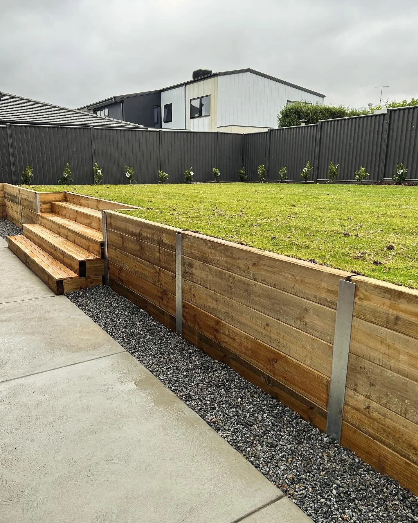 Wooden retaining wall with gravel, grass, and steps leading to a yard, backed by a fence and houses — MGP Landscaping in Geelong, VIC 