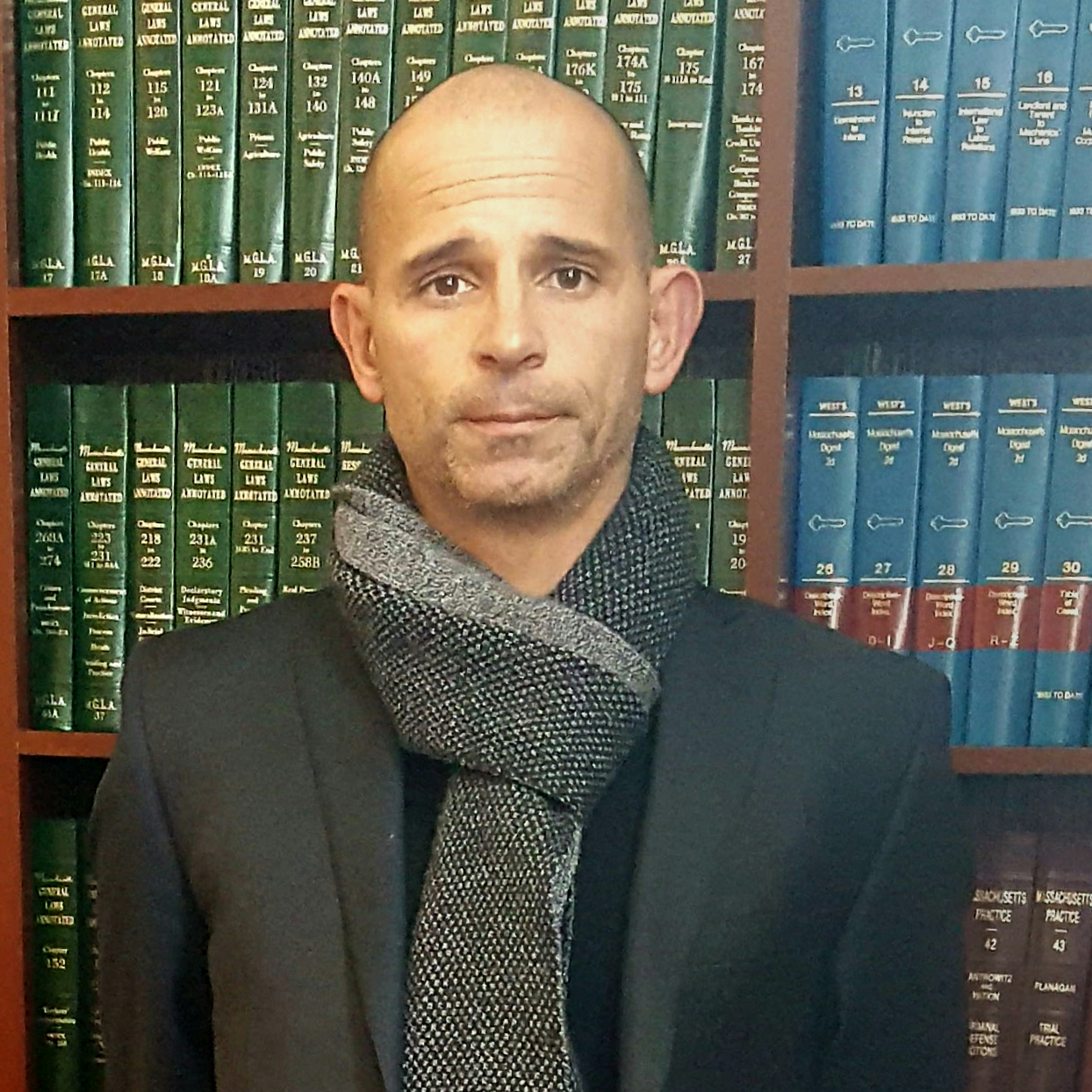 Man in dark suit and gray scarf standing before a bookshelf of green and blue law books.