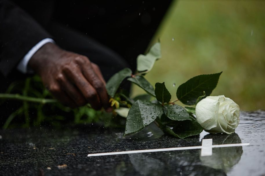 Person holding a white rose at a funeral; people in black attire are in the background. Person holding a white rose at a funeral; people in black attire are in the background.