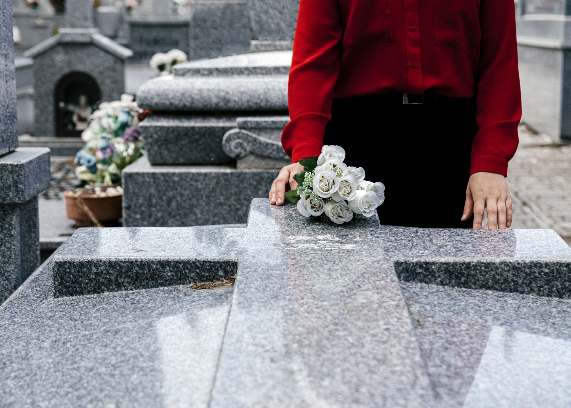 Person in red shirt and black pants places flowers on a gray stone cross in a cemetery. Person in red shirt and black pants places flowers on a gray stone cross in a cemetery.