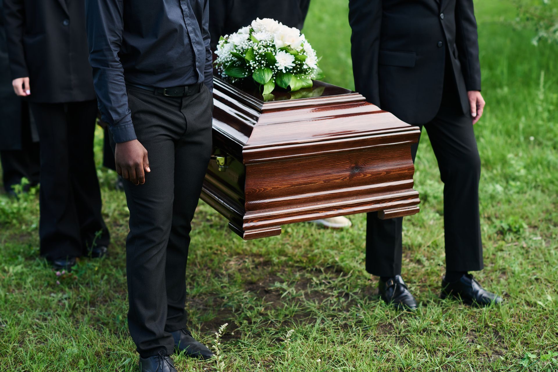 People in black clothing carrying a brown coffin with white flowers on green grass. People in black clothing carrying a brown coffin with white flowers on green grass.