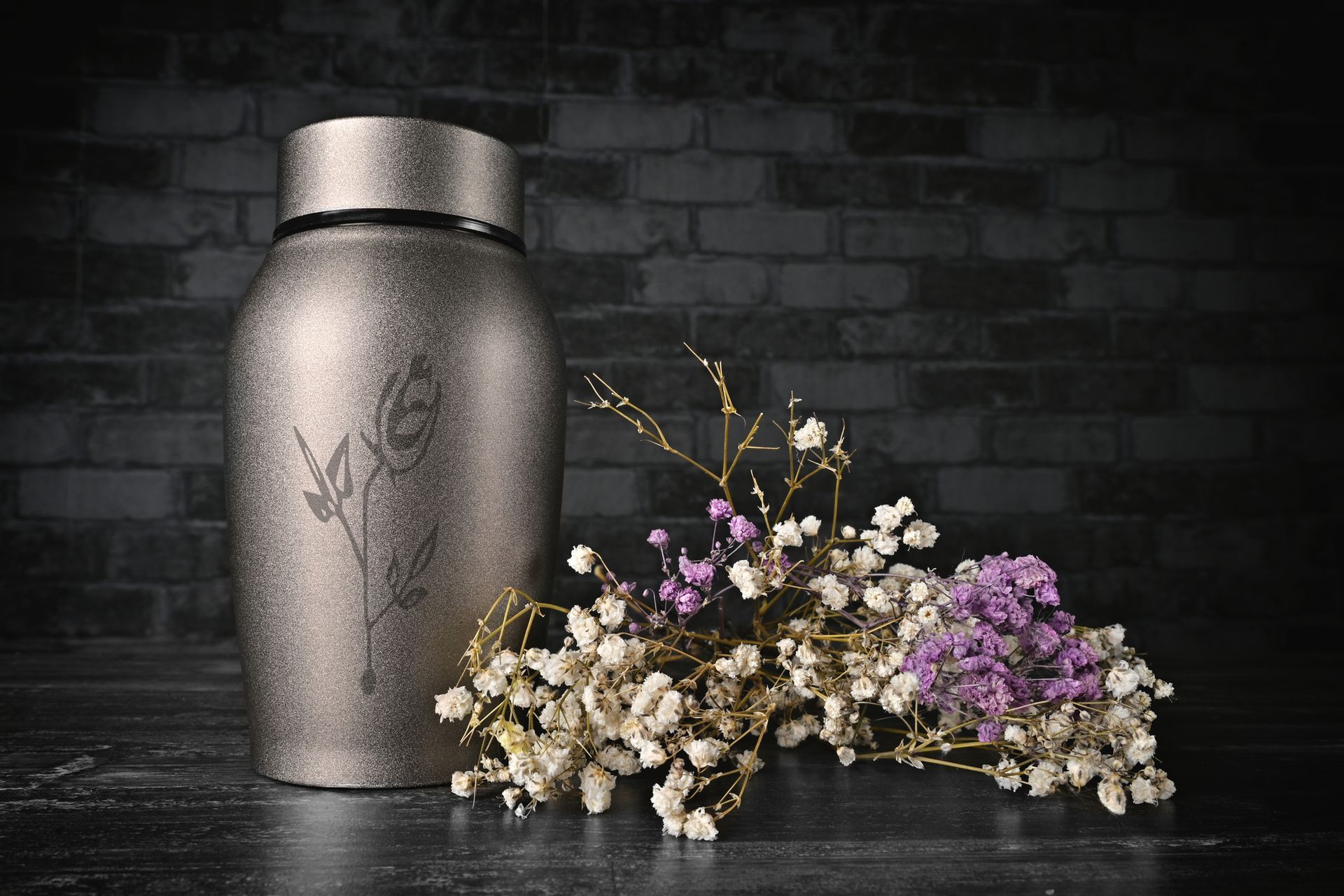 Silver urn with floral etching, beside a bouquet of baby's breath and purple flowers, against a brick wall. Silver urn with floral etching, beside a bouquet of baby's breath and purple flowers, against a brick wall.