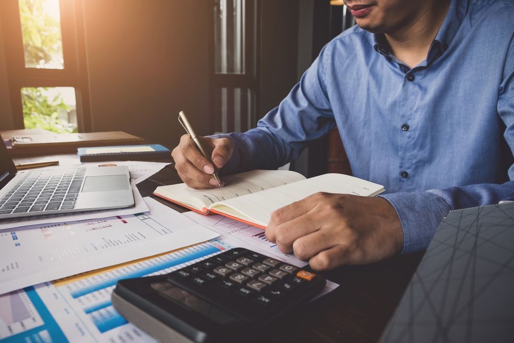 A Man Is Sitting At A Desk Using A Calculator And Writing In A Notebook — Emmel Finance in Townsville, QLD