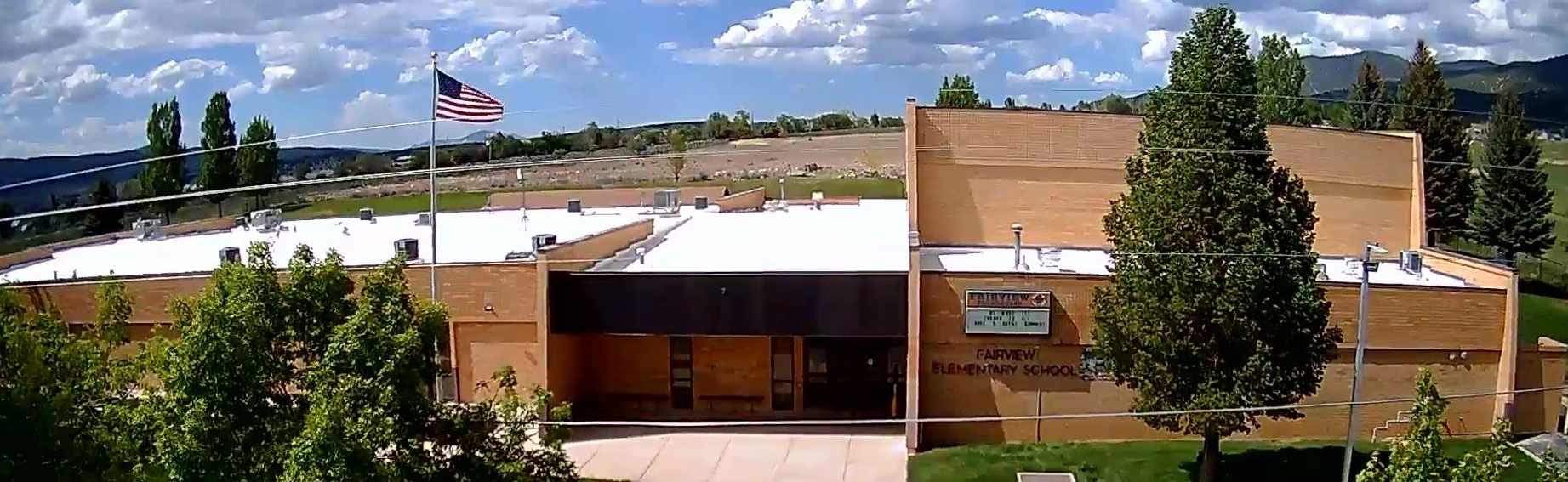 Exterior of a light brown building with white roof, trees, and flag on a sunny day.