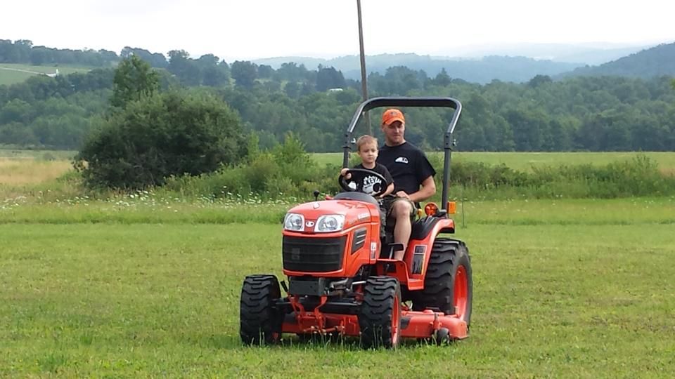 Man and child driving an orange tractor in a grassy field; mountains in the background.