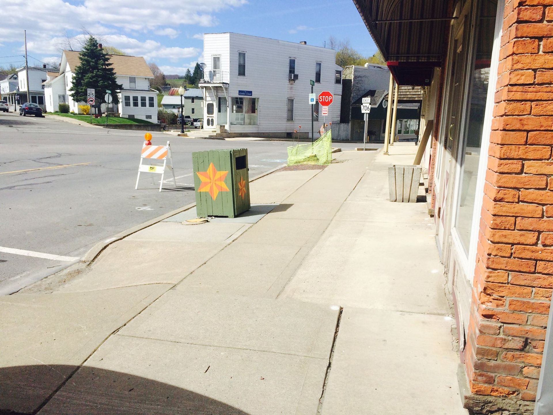 Sidewalk view on a sunny day. A green trash can with a star design is on the sidewalk near a street corner.