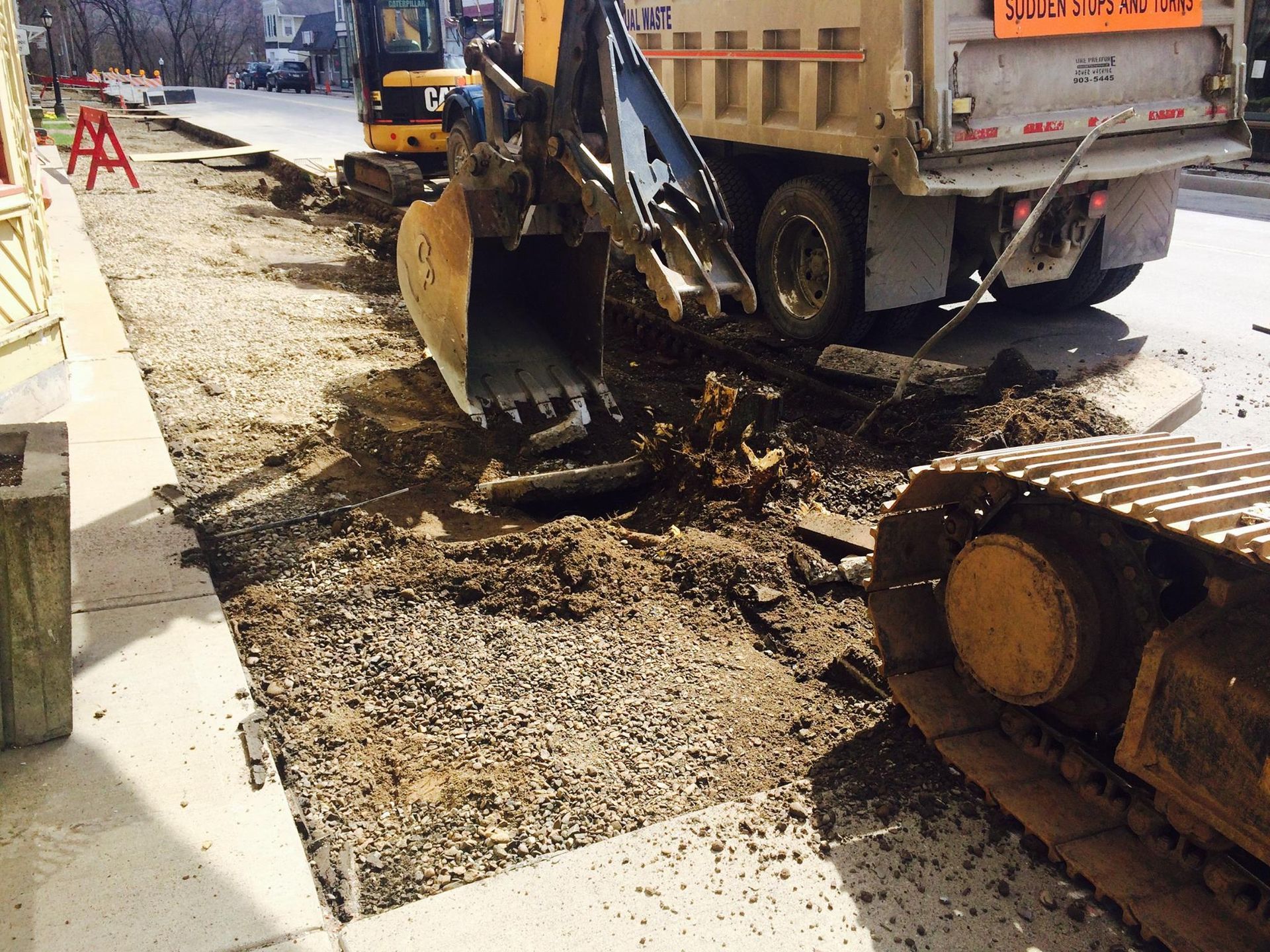Construction on a sidewalk: excavator loading debris into a dump truck, alongside a building.