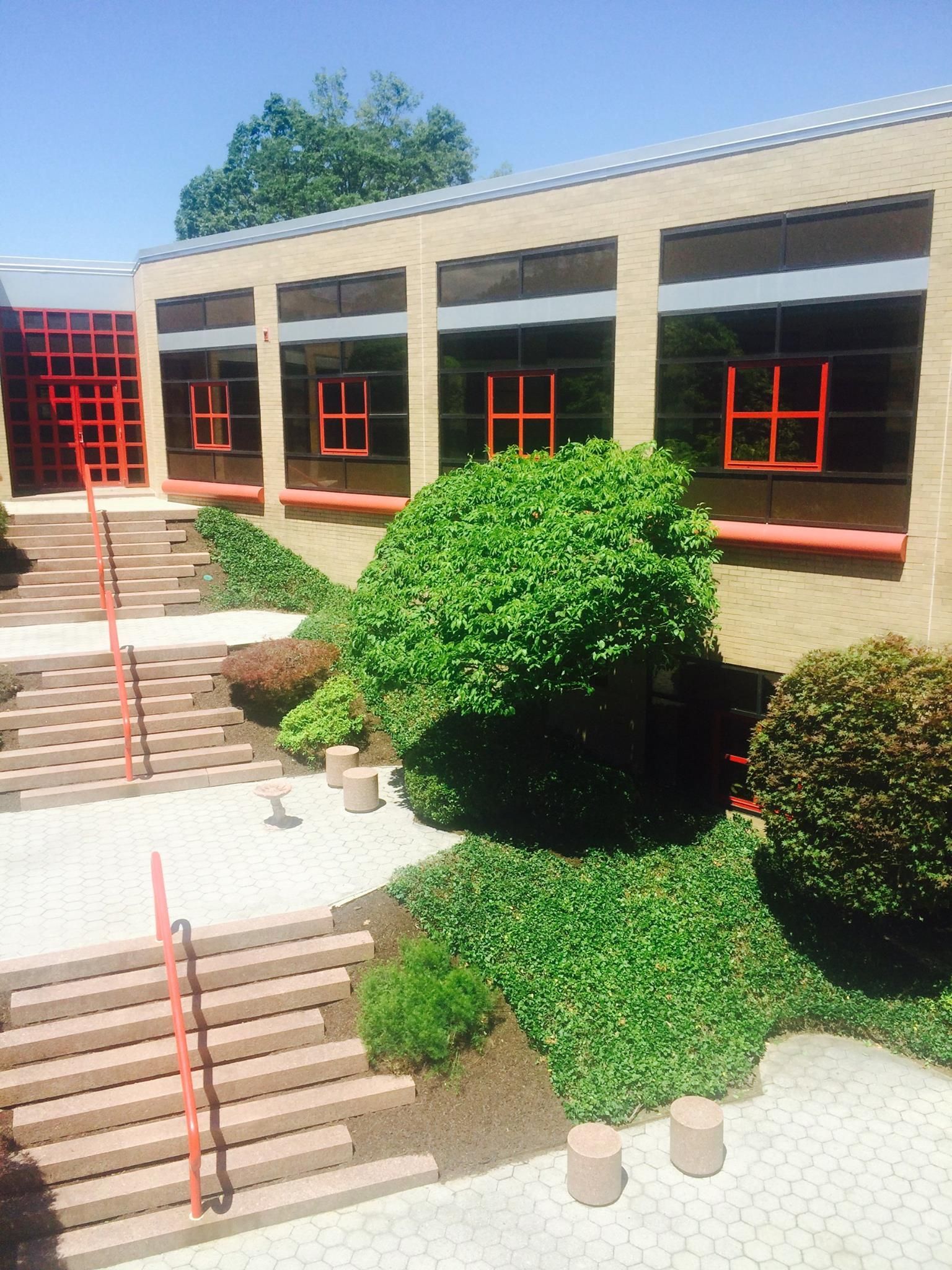 Courtyard with stairs, greenery, and a building with orange window frames and door. Sunny day.