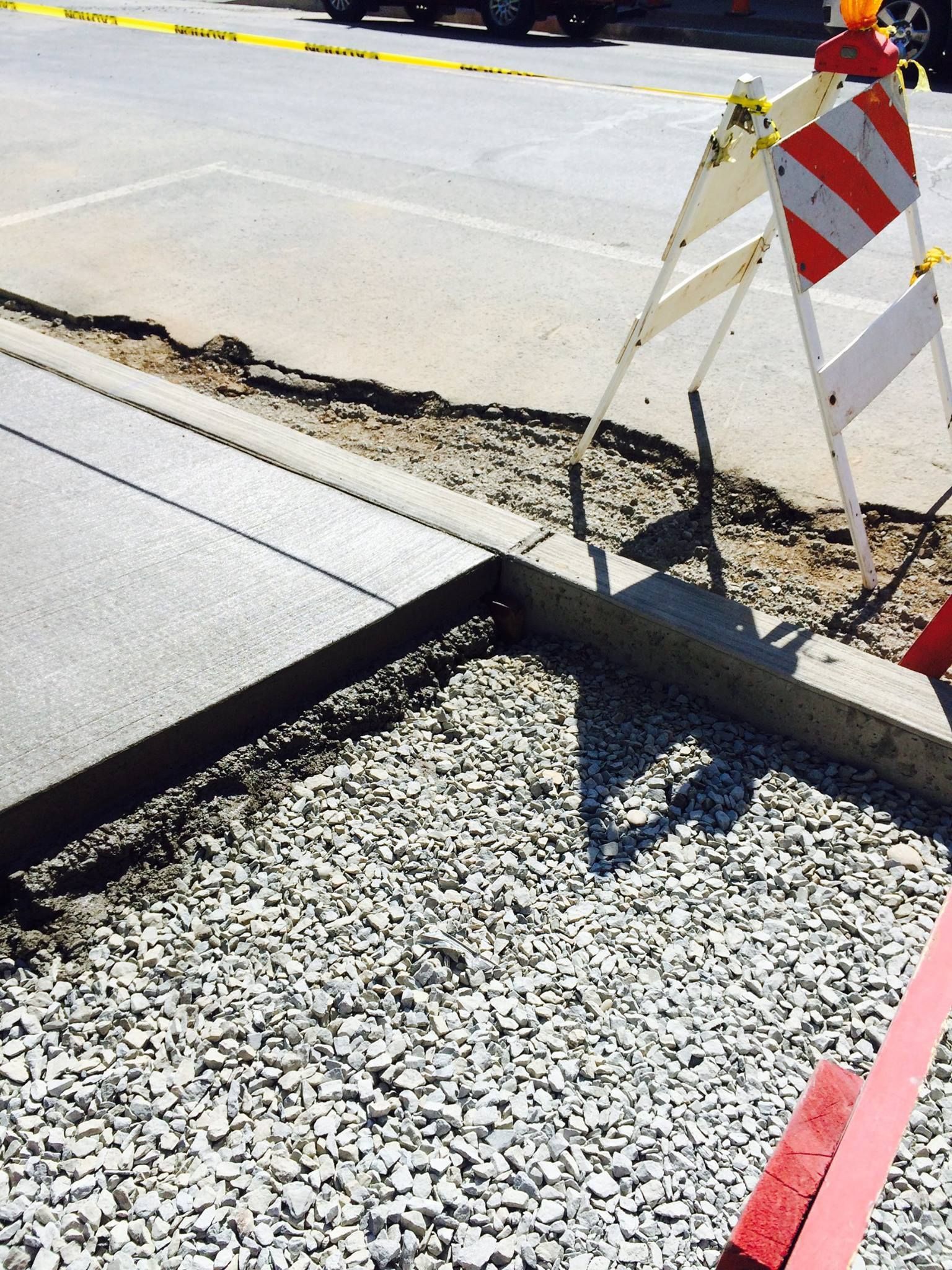 New concrete curb and sidewalk construction, gravel bed, safety barricade present.