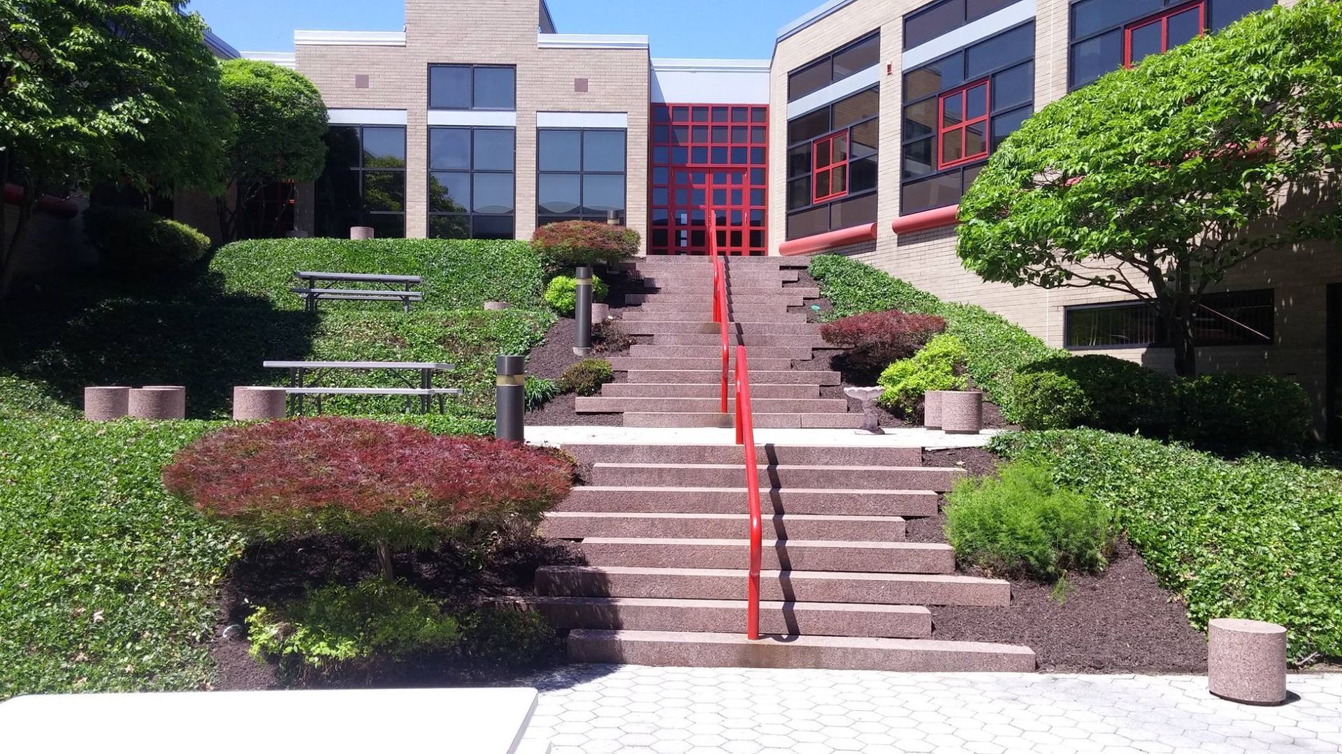 Staircase leading up to a building entrance; red handrails, bushes, benches on either side, sunny day.