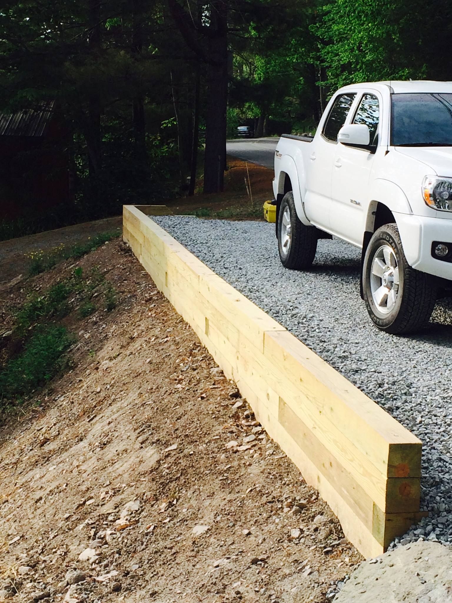 White pickup truck parked on gravel driveway next to a wooden retaining wall on a dirt embankment.