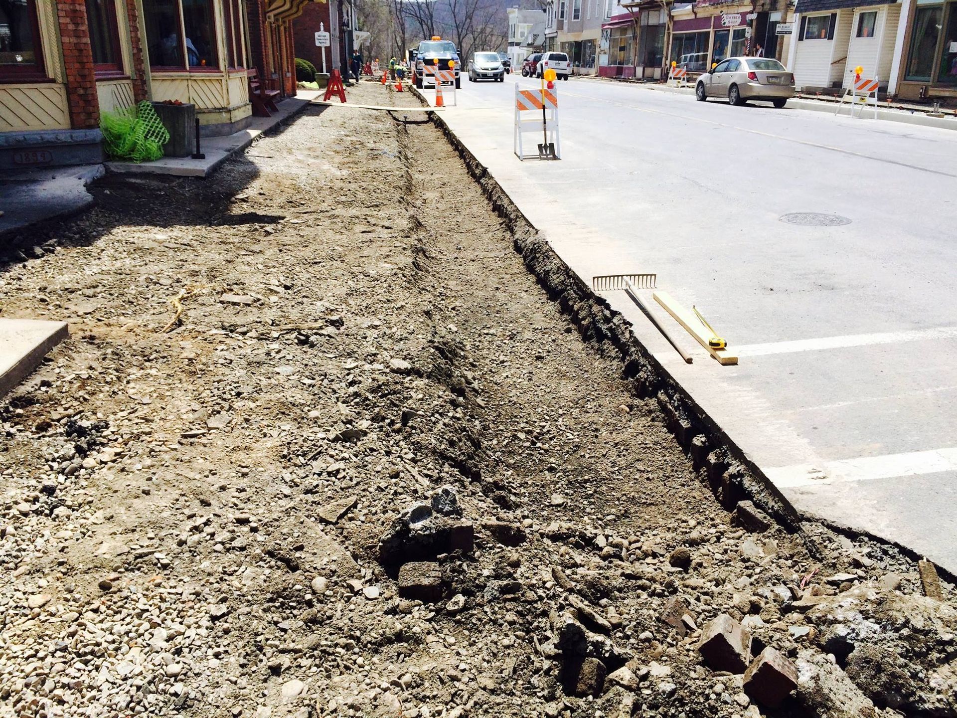Sidewalk under construction; dirt and debris in foreground, street and cars visible in the background.