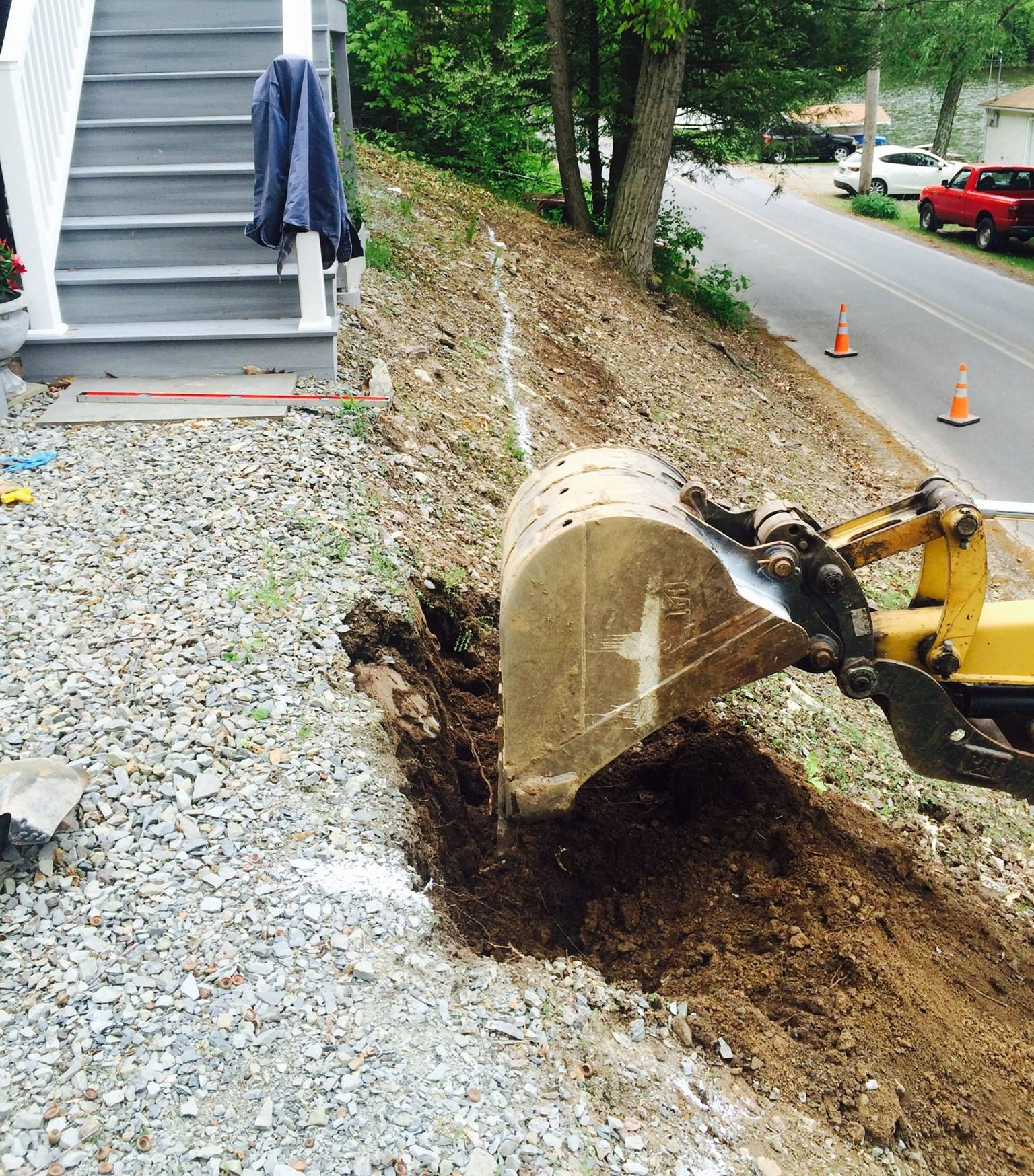 Yellow excavator digging a trench along a gravel driveway next to a steep hillside and stairs. Road in background.