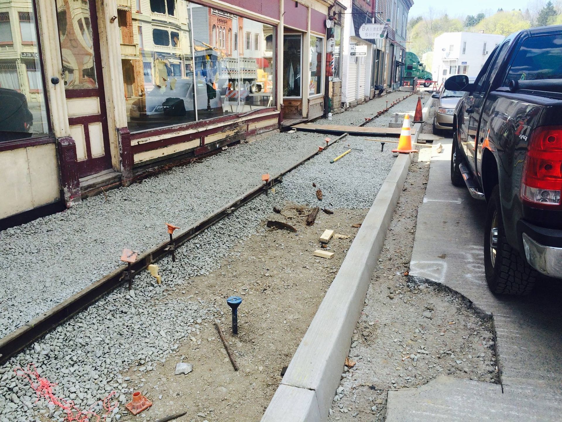 Sidewalk construction on a street with shops; gravel, concrete, tools visible. Car parked on right.