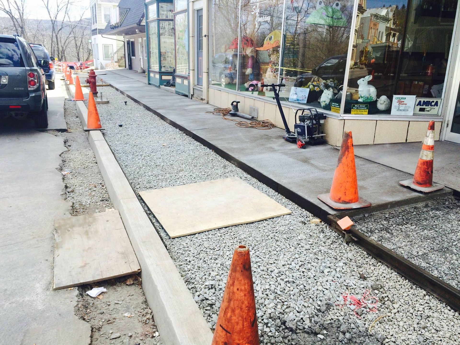Sidewalk construction with orange cones. Gravel and concrete work along a storefront.