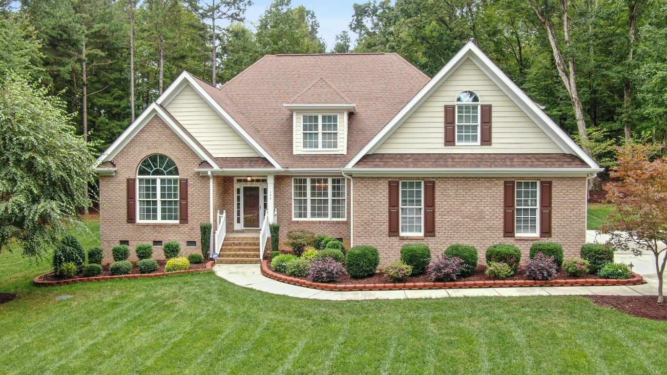 Brick house with brown roof and shutters, front yard with green grass, landscaping, and trees in background.