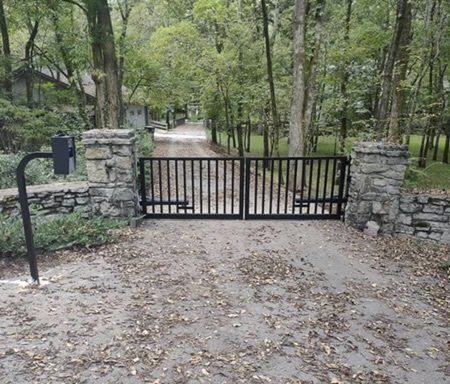 a black gate is surrounded by stone pillars on the side of a dirt road .