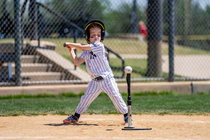 Young baseball batter in a white uniform swings at a tee on a sunny field.