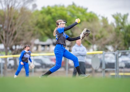 Softball player in blue uniform catches a ball midair on a grassy field while teammates watch behind her