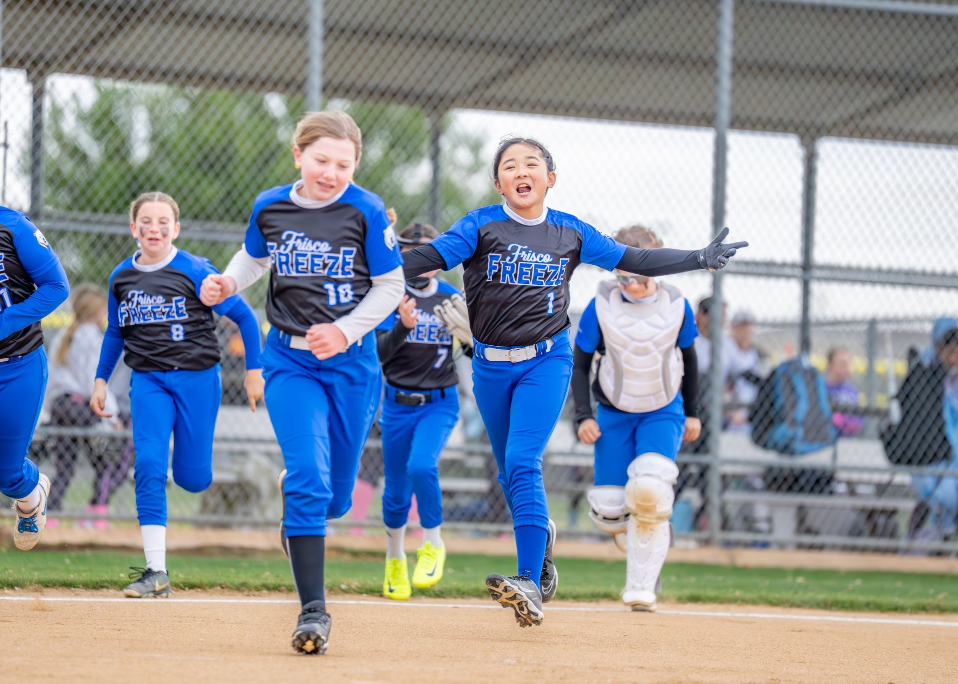 Youth softball players in blue uniforms sprinting toward home plate on a dirt field