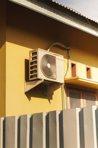 Wall-mounted air conditioner units on a yellow building above a gray metal fence.