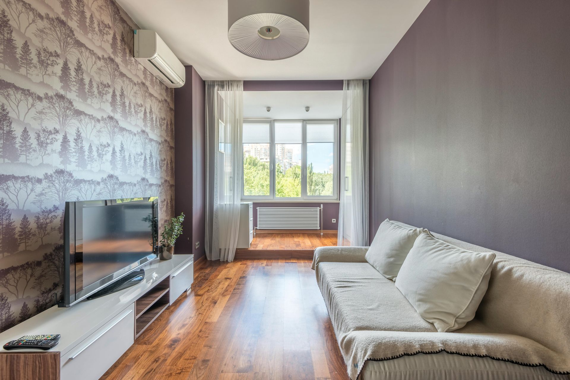 Bright living room with a sofa, TV, hardwood floors, patterned accent wall, and large window with curtains