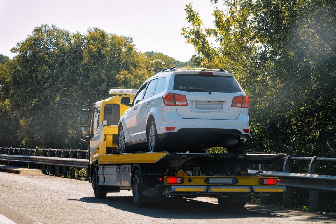 White car being towed on a yellow flatbed tow truck, on a road. Trees in the background.