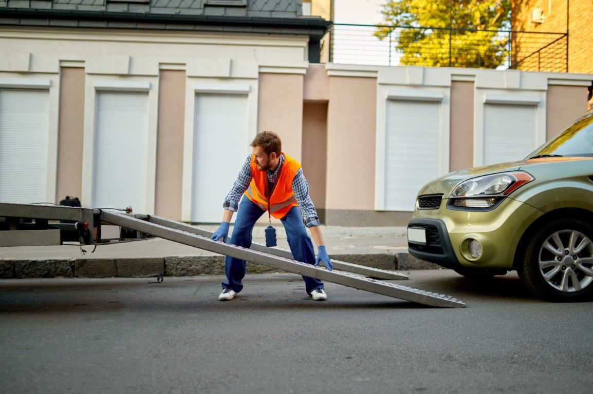 Man in orange vest struggling to set up a ramp to tow a green car on a city street.