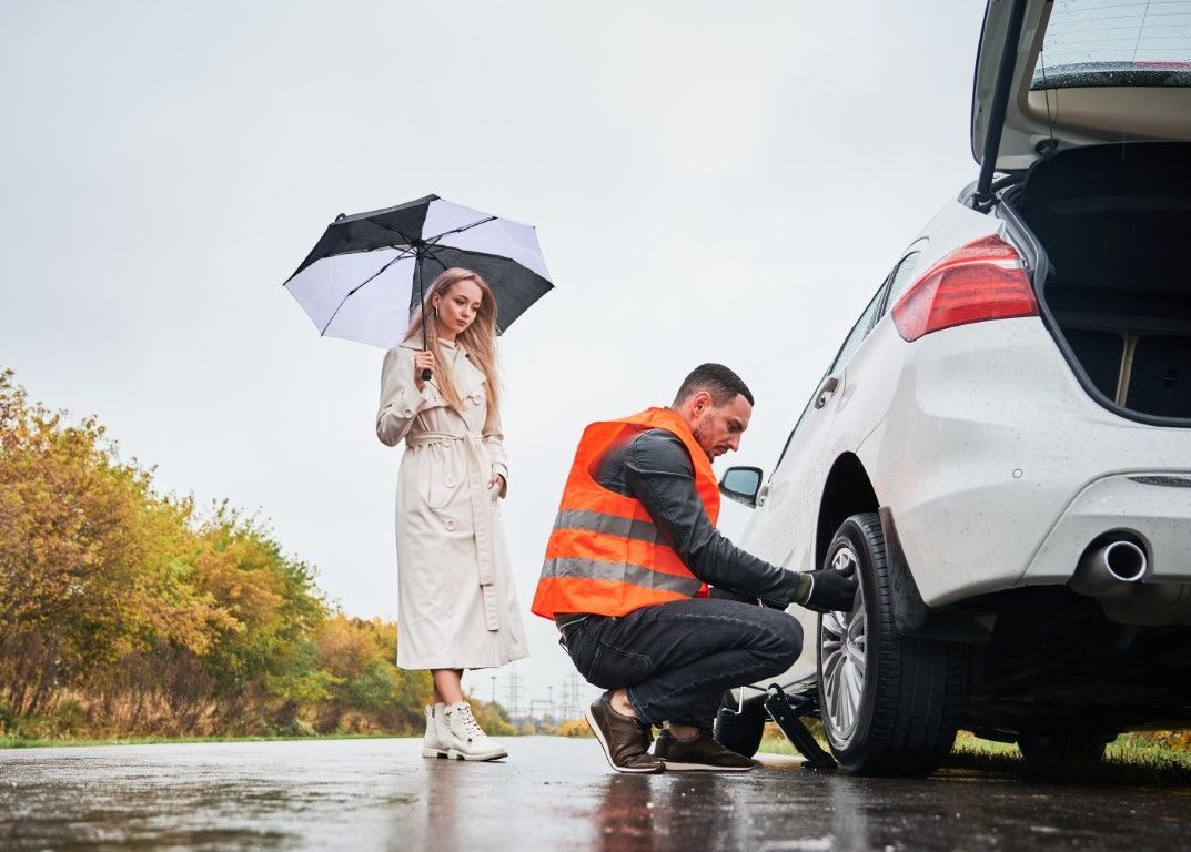 Man changing a tire on a white car in the rain; woman holding an umbrella watches.