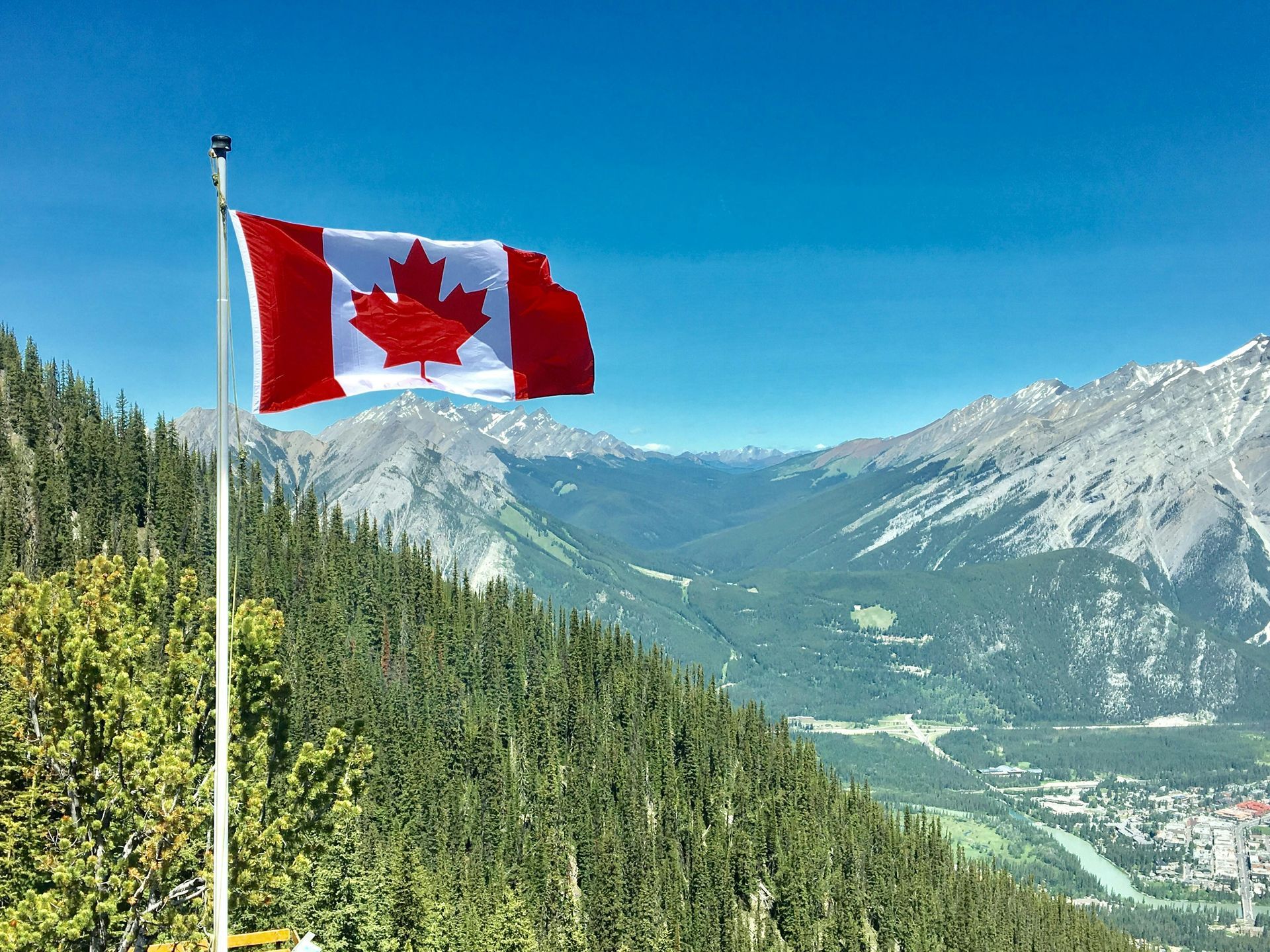 Canadian flag waving in the wind against a mountain vista, blue sky, and green forest.