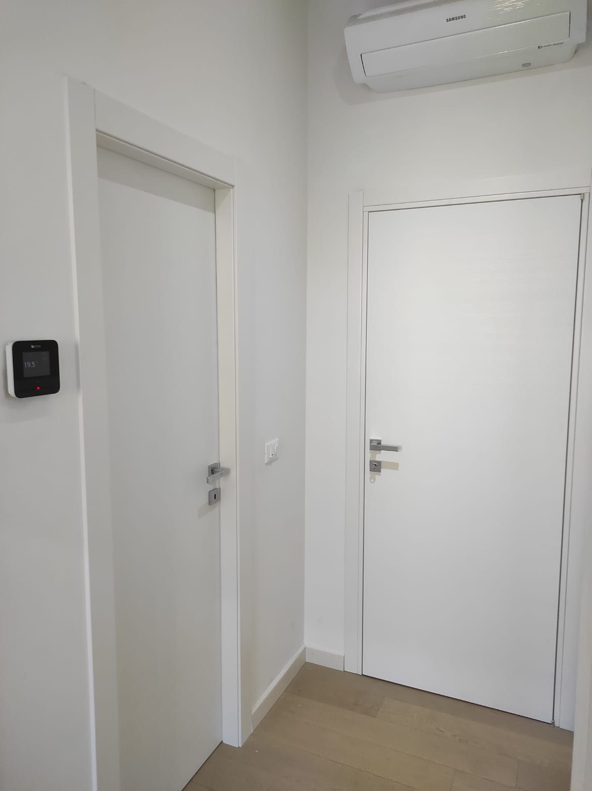 Due porte interne bianche in un corridoio dai toni neutri, con un piccolo termostato digitale sulla parete sinistra e un condizionatore d'aria sopra.