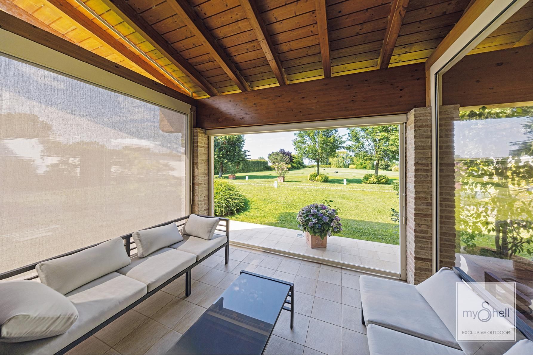 Un patio esterno coperto con divani dai colori chiari, un tavolino da caffè e vista su un rigoglioso prato verde.