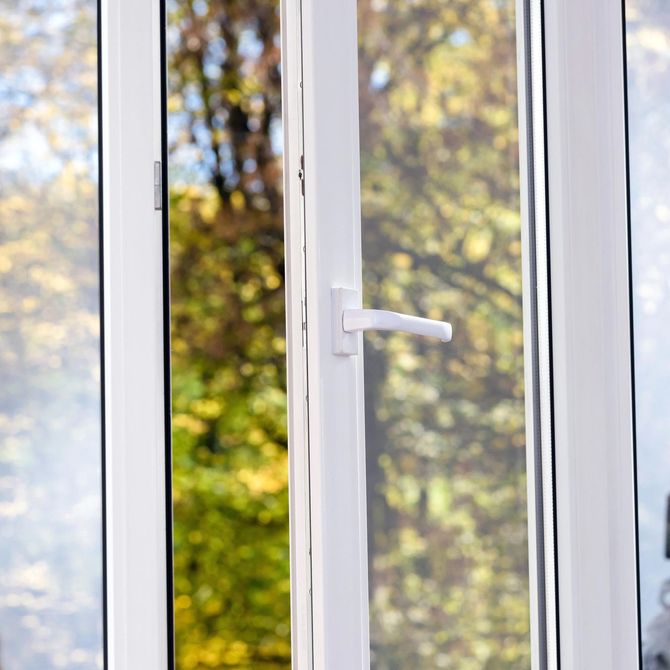 Un telaio di finestra in PVC bianco con maniglia è leggermente socchiuso, lasciando intravedere, in modo sfocato, gli alberi dai colori autunnali all'esterno.