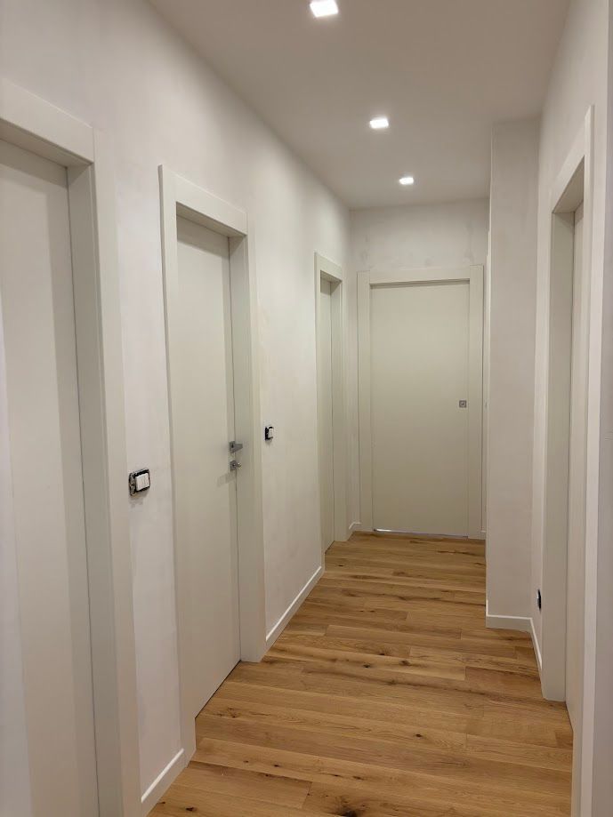 Un corridoio con pareti bianche e pavimento in legno chiaro, caratterizzato da diverse porte bianche che conducono alle stanze e faretti a incasso sul soffitto.