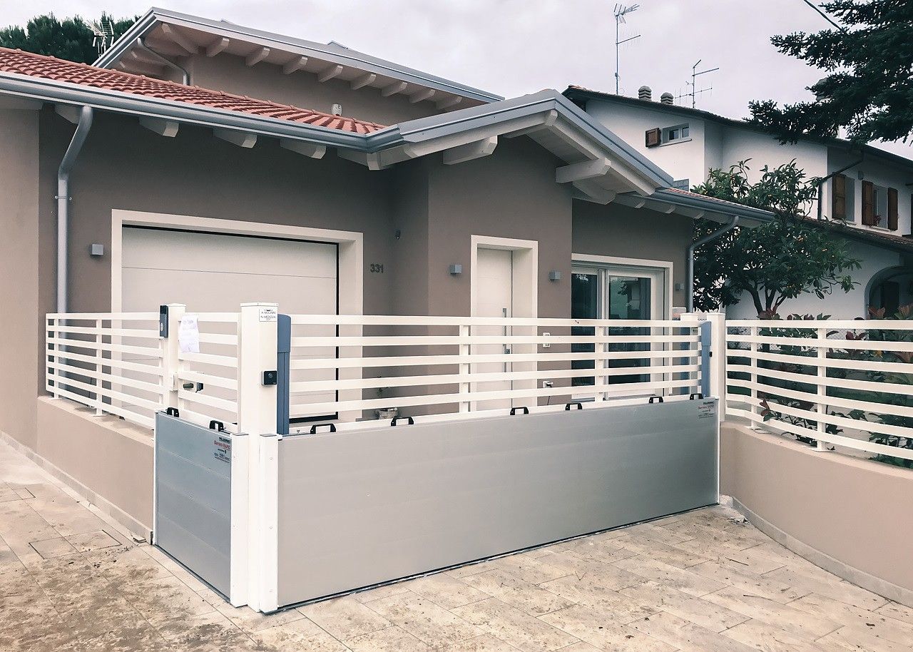 Una casa grigia e moderna con una recinzione bianca e una barriera metallica antiallagamento installata sul vialetto d'accesso.