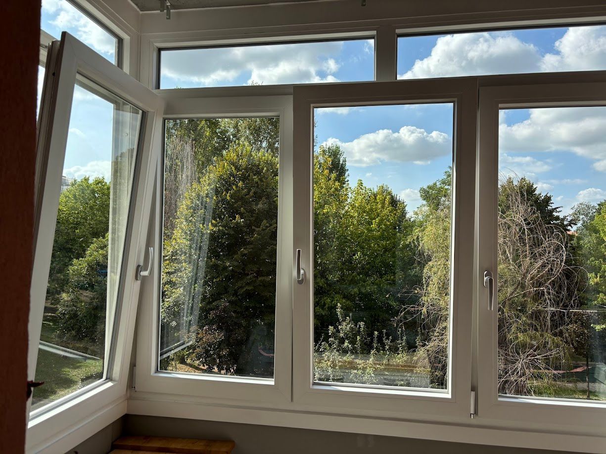 Una finestra con cornice bianca e diverse ante, una delle quali aperta, che si affaccia su un paesaggio luminoso, verde e soleggiato.