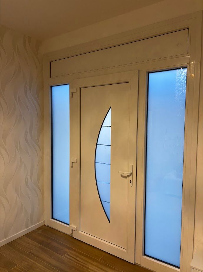 Una porta d'ingresso bianca con un pannello di vetro curvo e finestre laterali, su uno sfondo di carta da parati a motivi geometrici e pavimento in legno.