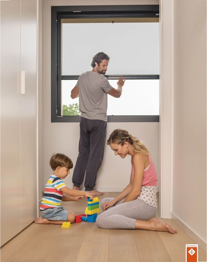 Una persona sistema una zanzariera mentre un bambino e un'altra persona giocano sul pavimento con dei colorati mattoncini di plastica.