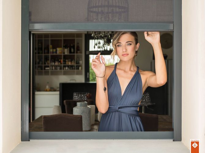 Una persona con un abito blu a portafoglio posa dietro una finestra aperta con una zanzariera grigia, in un interno con un arredamento domestico sfocato.