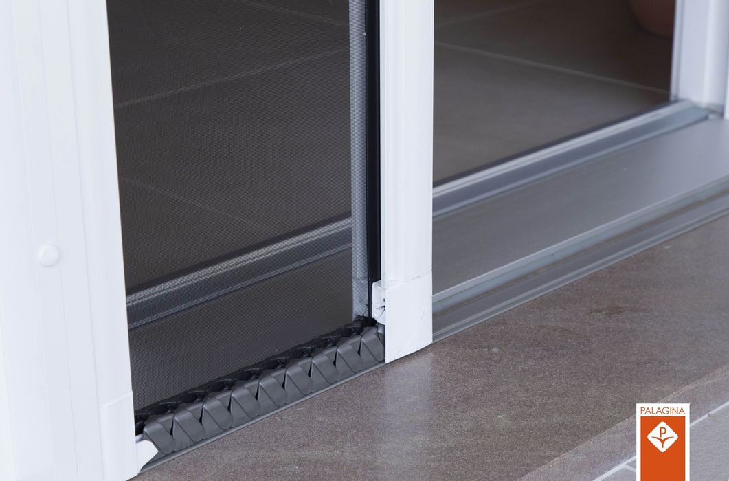 Una porta a zanzariera a scomparsa e plissettata, installata in un telaio bianco su un pavimento grigio.