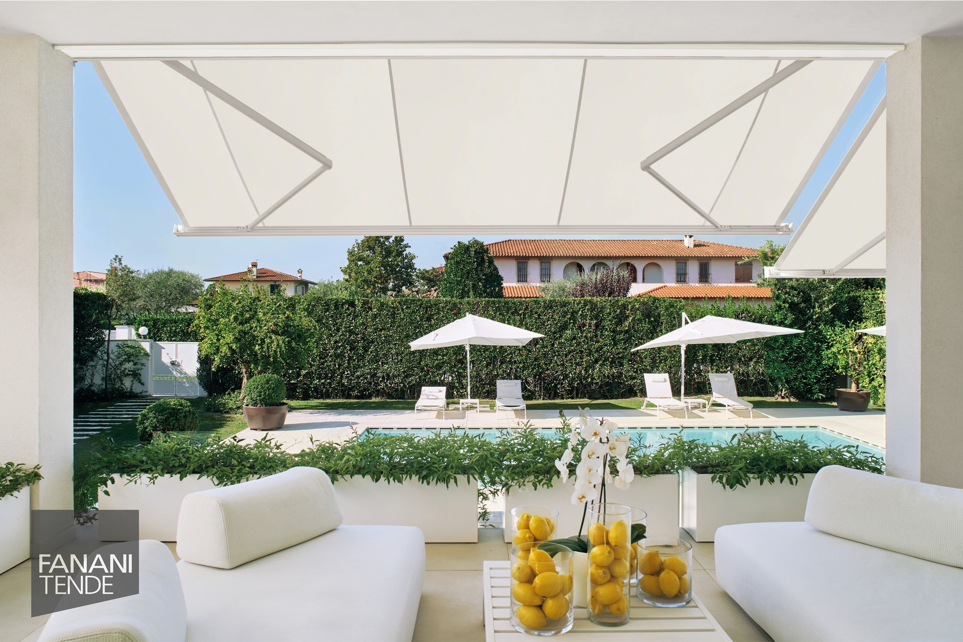 Una tenda da sole retrattile bianca ombreggia un patio esterno con divani bianchi, un tavolo con limoni e una piscina sullo sfondo.