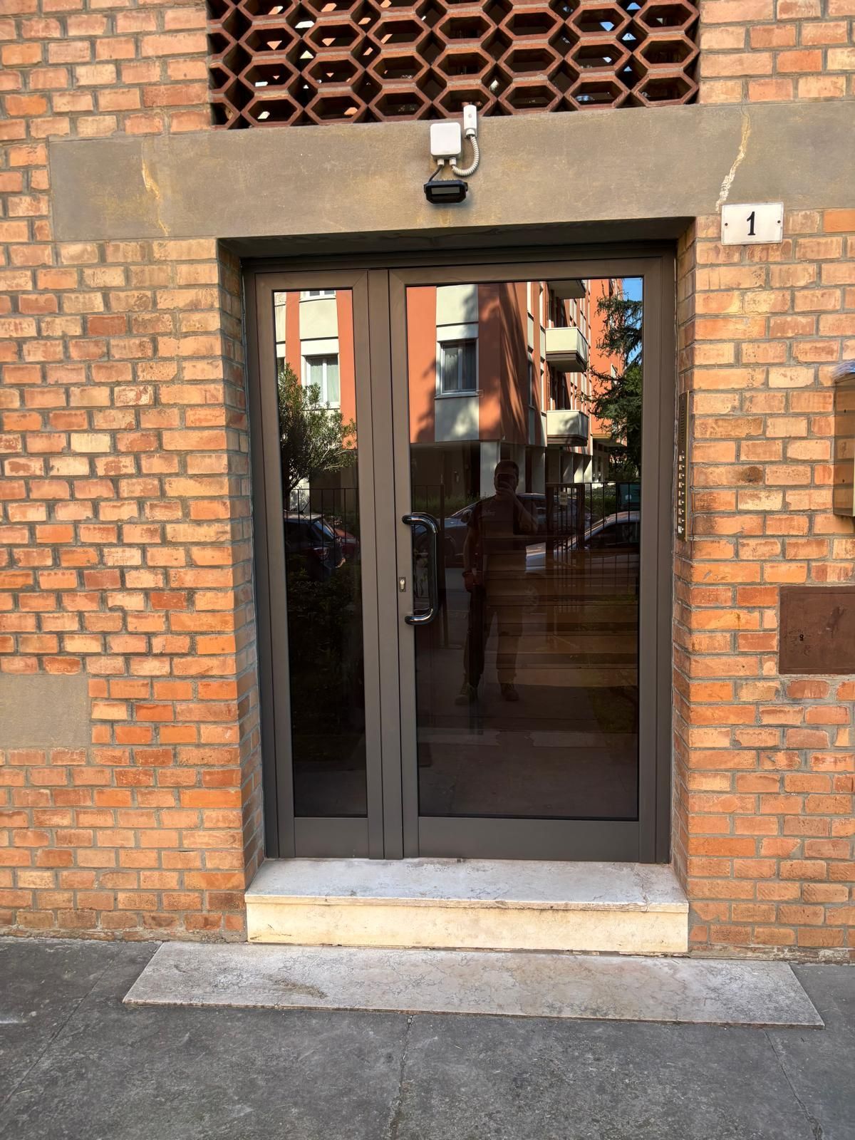 Una persona percorre una stretta strada acciottolata, passando davanti a un edificio giallo con due ingressi incorniciati di scuro e piante in vaso.