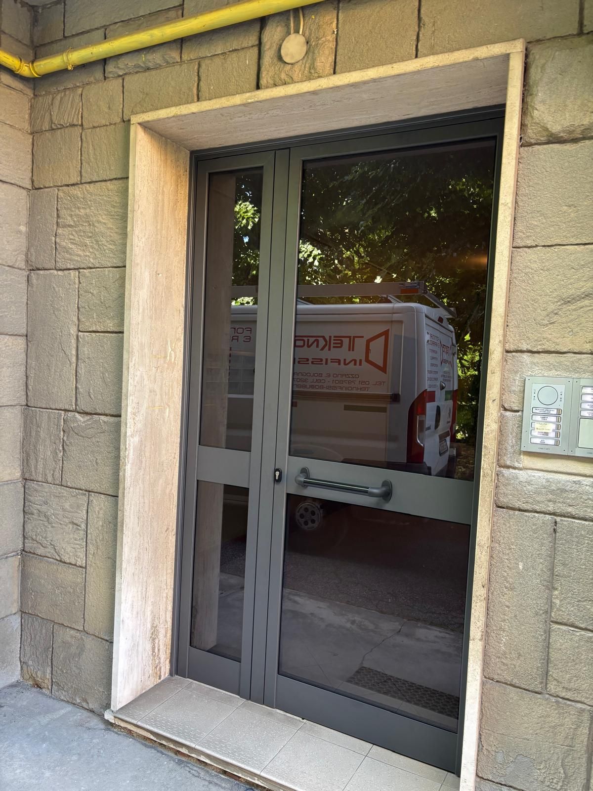 Una facciata color beige con due porte d'ingresso in legno scuro, pannelli laterali in vetro e una siepe ornamentale in vaso ai lati di ciascun ingresso.