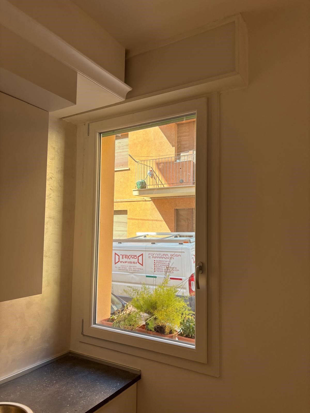 Una finestra rettangolare con cornice bianca che si affaccia su una strada soleggiata con un furgone bianco parcheggiato e un piccolo balcone.