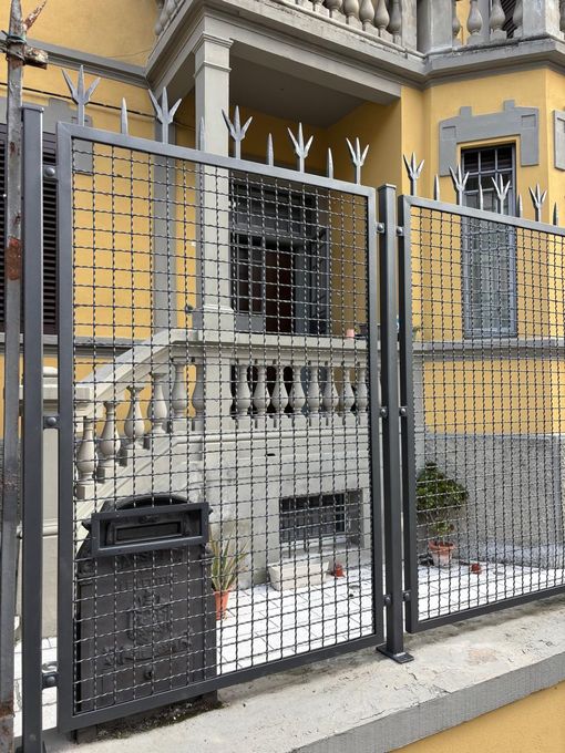 Un cancello di metallo con sbarre superiori appuntite si erge davanti a un edificio giallo con scale in pietra e una cassetta delle lettere nera.