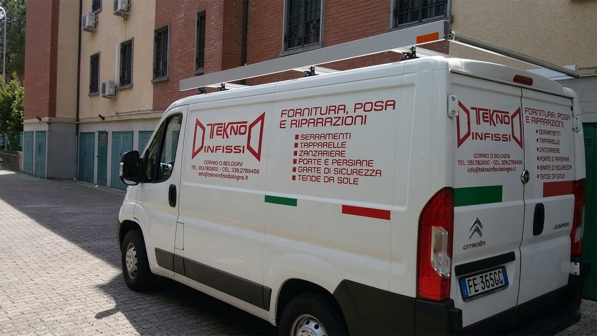 Un furgone bianco adibito a servizio, parcheggiato in un piazzale asfaltato, mostra il logo aziendale rosso, scritte e una bandiera italiana sul fianco.